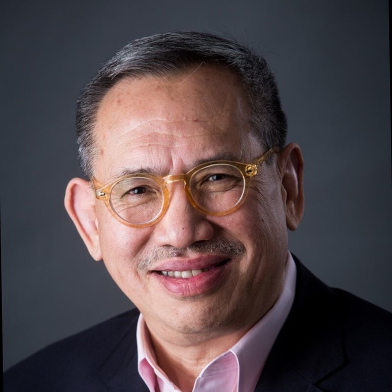 A man with short grey and black hair wears translucent yellow glasses, a black suit jacket, and light pink button down.