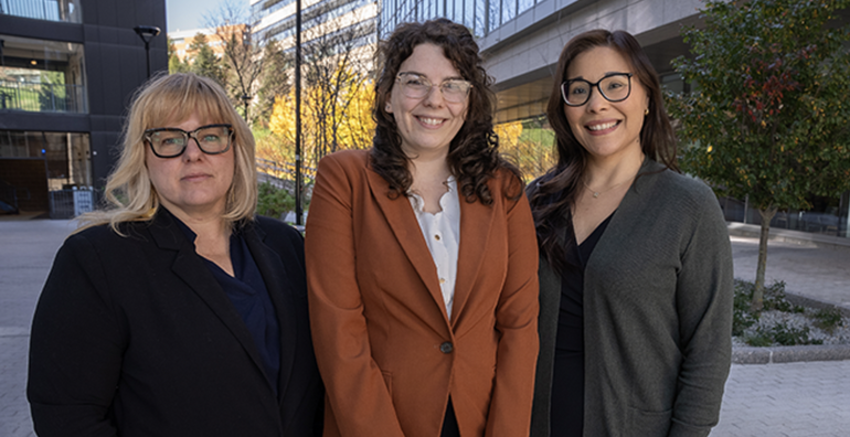 Three women stand side-by-side outside university buildings. The woman on the left has blonde hair with bangs, wears black glasses, a black suit jacket, and navy top. The woman in the middle has curly brown hair, wears clear glasses, a burnt orange suit jacket, and white top. The woman on the right has long brown hair and wears black glasses, a grey cardigan, and black top.