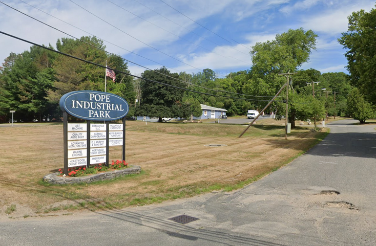 A sign for an industrial park with smaller signs underneath listing tenants sits in an open field next to a driveway