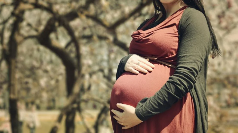 A woman wears a reddish-brown top and green cardigan as she holds her baby bump while standing outside in front of trees.