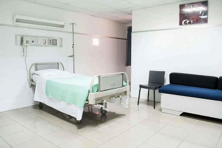 A hospital bed with white sheets and a mint green blanket sits inside a white hospital room with white tiled floors and a couch with blue cushions against the wall.