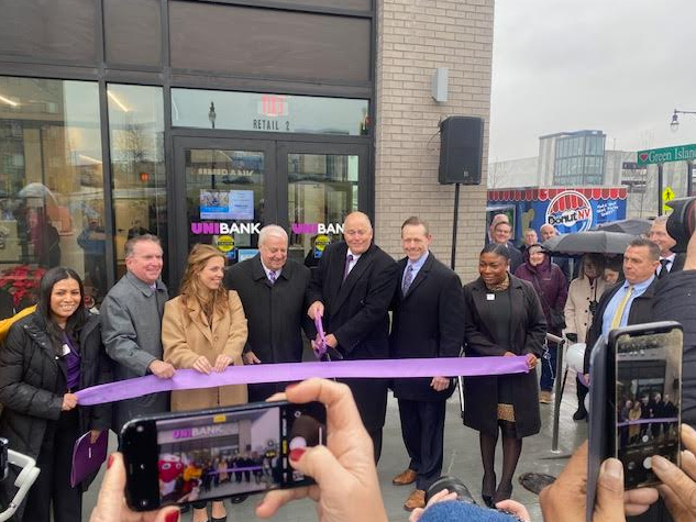 A group of people stand outside of a bank and behind a large purple ribbon they're holding while one man holds a pair of purple scissors to cut it.