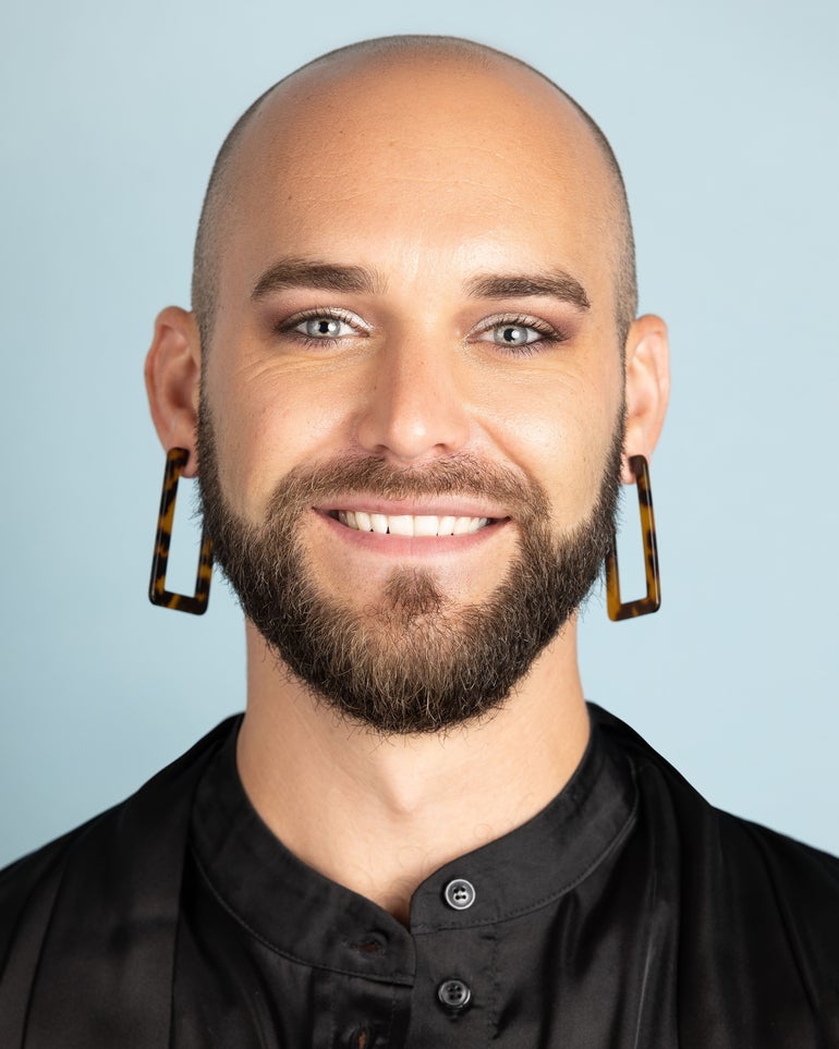 A person with a beard, large square earrings, and a black shirt smiles at the camera.