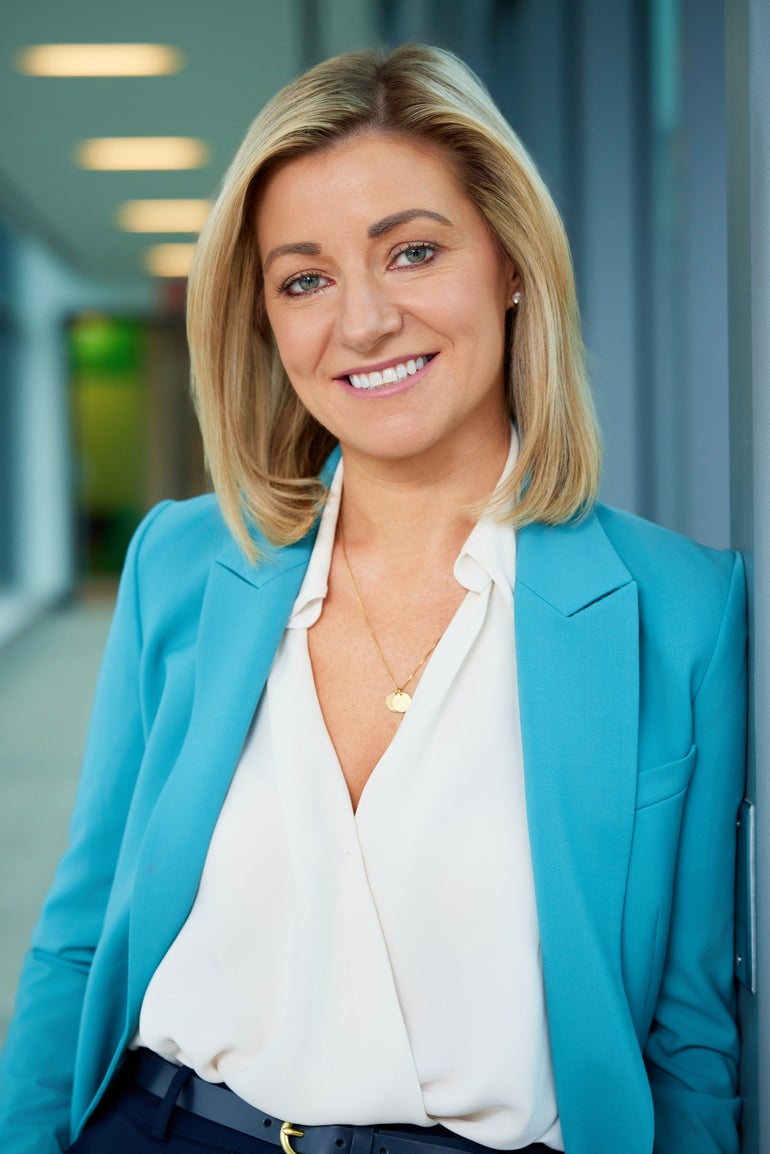 A woman with shoulder length blonde hair wears a light blue blazer, a white v-neck top, and a gold necklace while leaning against a wall.