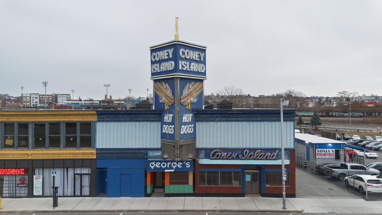 One grey and yellow building attached to a light and dark blue building with a large sign about it reading "CONEY ISLAND" sits in back of a grey sidewalk with a parking lot on the right
