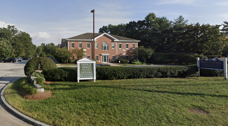 A large brick building sits behind a parking lot behind a grassy area with bushes.