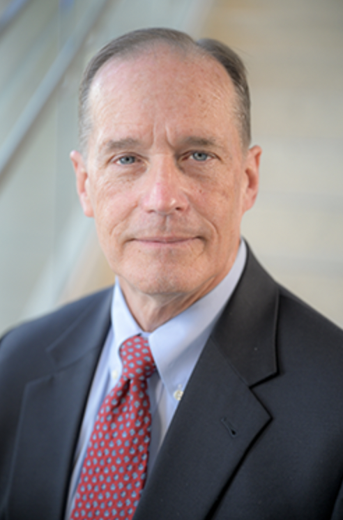 A man with grey hair wears a black suit jacket, a light blue button down, and a red patterned tie.