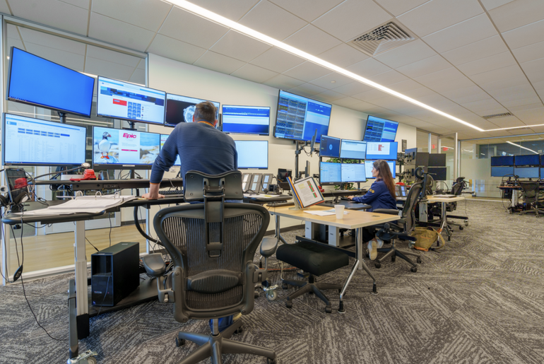 Two desks with multiple computer monitors with people standing/sitting in front of them