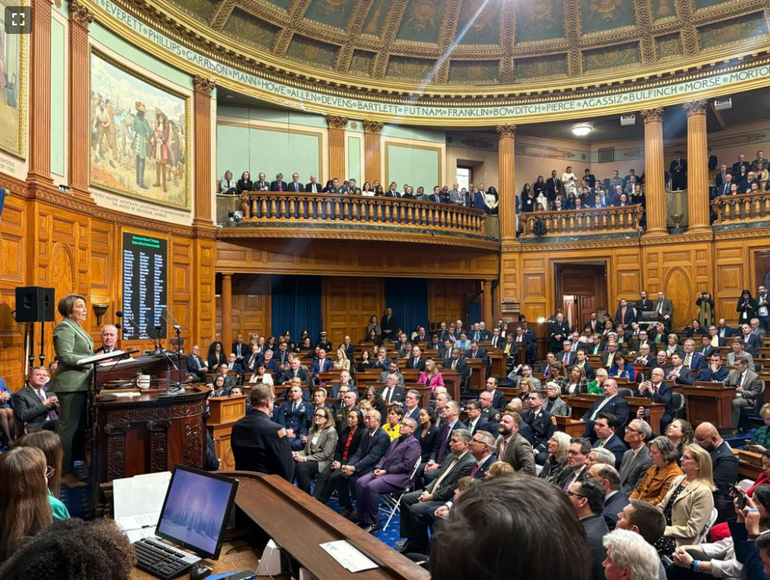 A woman stands at a podium in a crowded legislative chamber