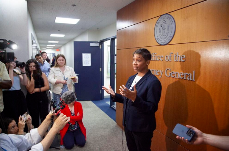 A woman in a blue suit stands talking to reporters in a hallway with wood paneling behind her.