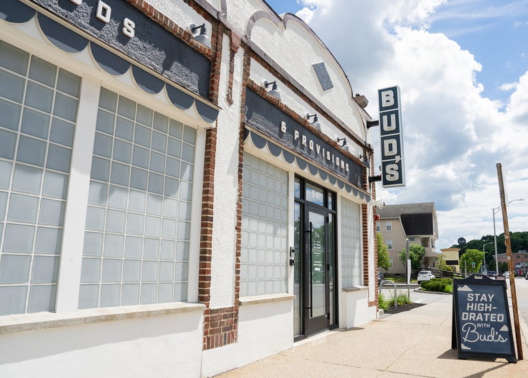 Outside a white building with a black door and a sign reading "BUD'S" sits behind a beige sidewalk.