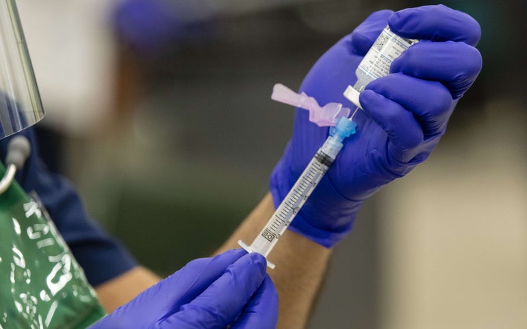 Hands wearing purple gloves hold a syringe as they draw a vaccine out of a container.