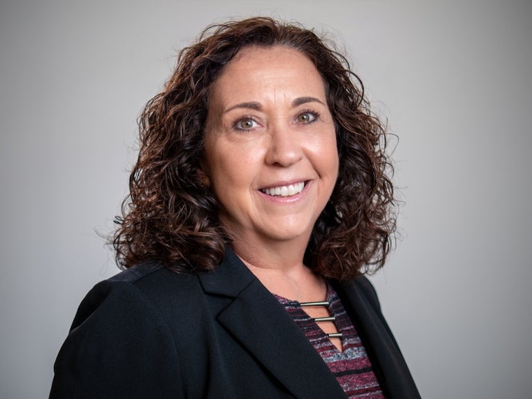A woman with shoulder length brown curly hair wears a black blazer and a purple, grey, and black patterned top.