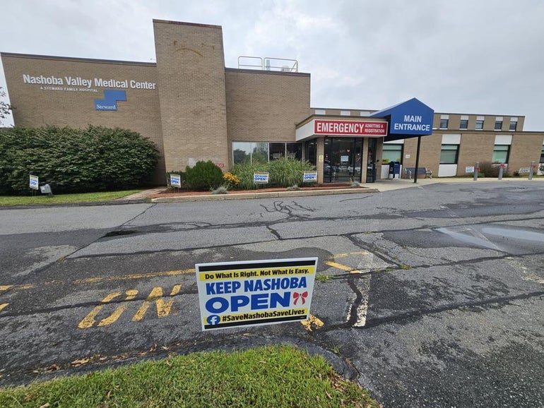 A brick emergency room sits behind a large driveway with a sign on it reading "Keep Nashoba Open".