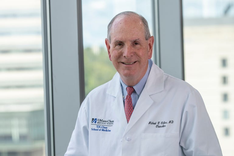 A man with grey hair wears a white lab coat, a light blue button down, and a red patterned tie.