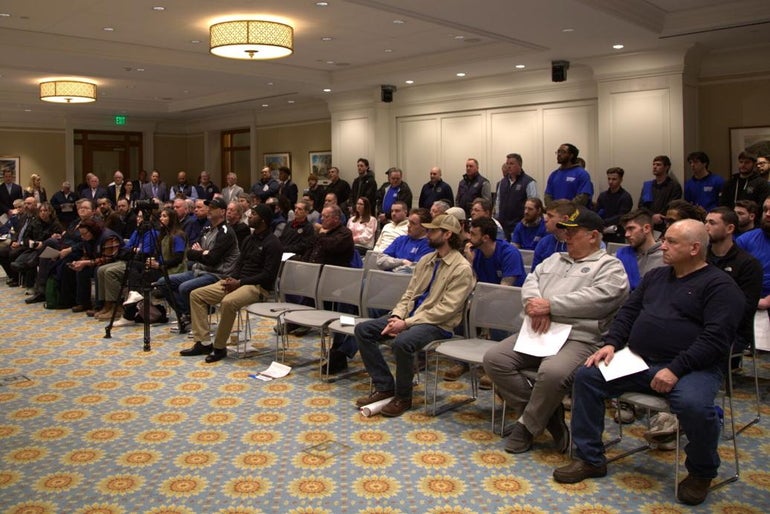 A large group of mostly men sit on chairs in a big room.