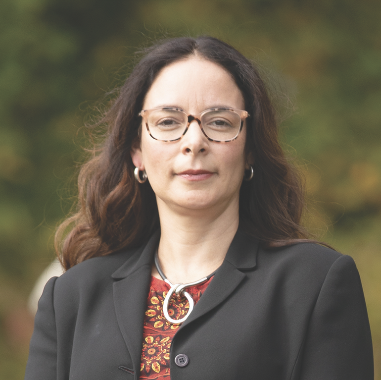 A woman with long brown hair wears glasses, a black blazer, and red patterned top.