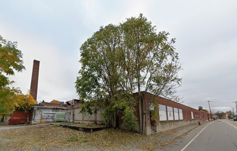 A one-story warehouse buildings with trees growing from the edge of the building and leafs scattered across a disused parking lot