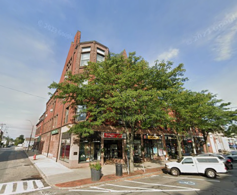Four-story brick building with first floor retail sits behind trees in a suburban downtown. 