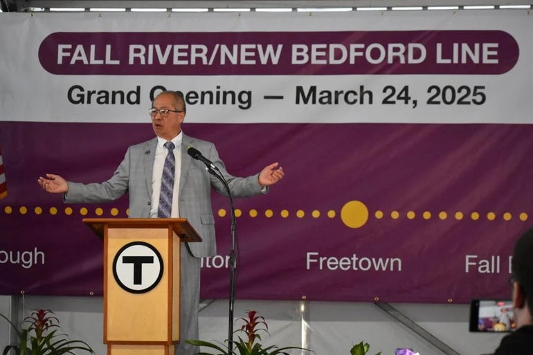 A man wearing a gray suit and purple tie stands behind a podium with the MBTA logo on it.