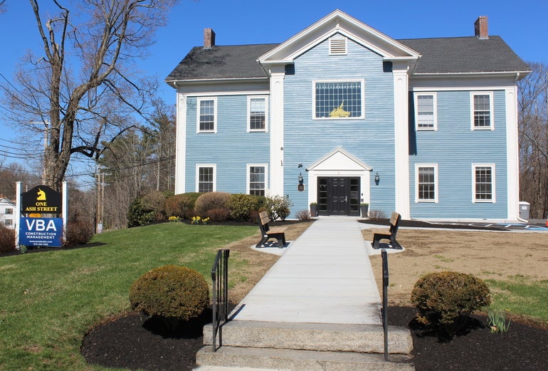 A two-story building resembling a house, with a paved path and benches in front.