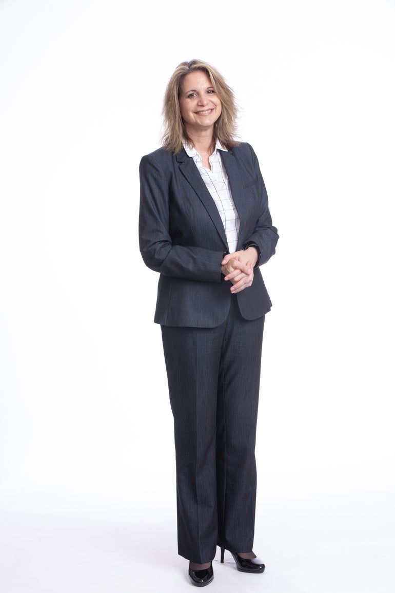 A woman with shoulder-length blonde hair stands with her hands together while wears a grey suit, a white button down, and black heels.