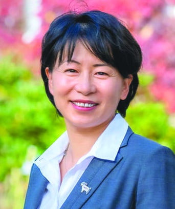 Headshot of a woman with short black hair wearing a blue suit jacket an white button down while standing in front of colorful leaves