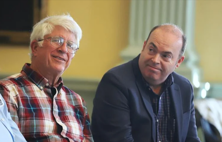 A man with glasses and another man sits next to one another. 