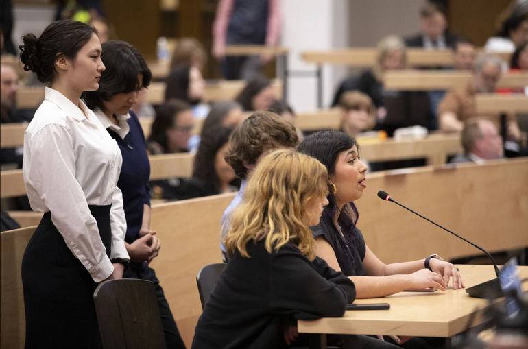 A group of five students stand and sit around a table with a microphone