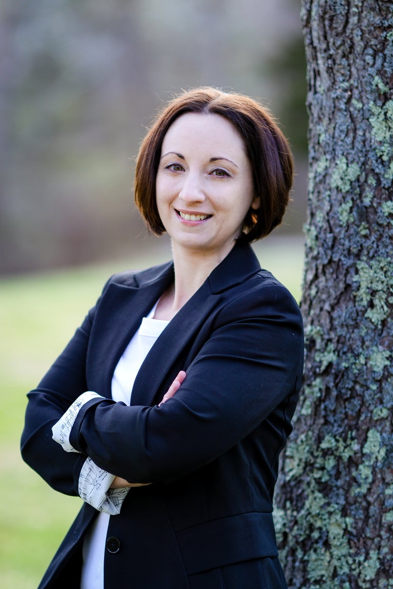 A woman with a short brown bob wears a black suit jacket, standing with her arms crossed in front of a tree.