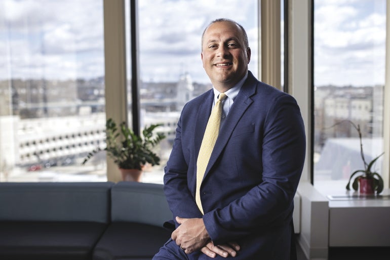 A man sits on a couch with his hands together wearing a blue suit in front of large office windows.
