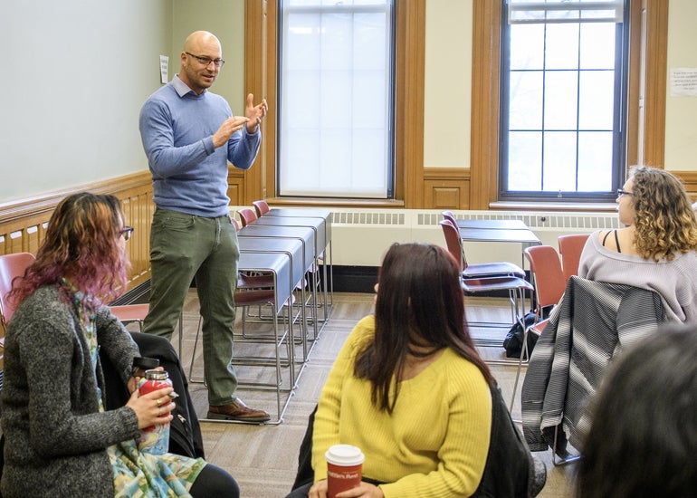 A mean wearing a blue sweater and green khakis stands inside a college classroom teaching to students seated around the classroom.