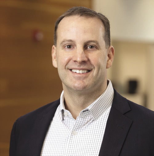 A man with short light brown hair wears a black blazer and da white checkered button down.