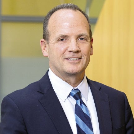 A man wears a dark blue suit jacket, a white button down shirt, and a dark and light blue striped tie.