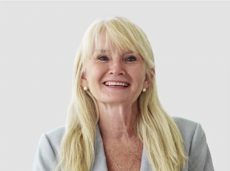 A woman with long, straight blonde hair and bangs wears a light grey blazer.