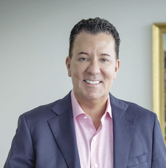 A man with short dark hair wears a dark blue suit jacket and pink and white checkered button down.