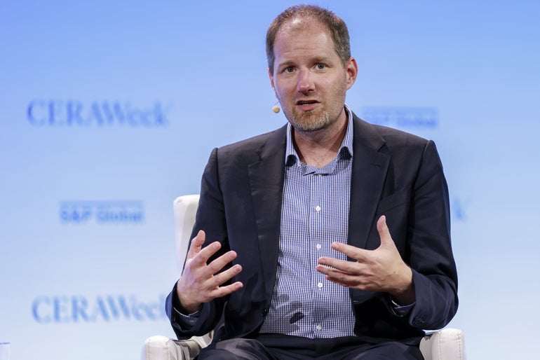 Man in suit with blue checkered dress shirt sits in a chair while speaking