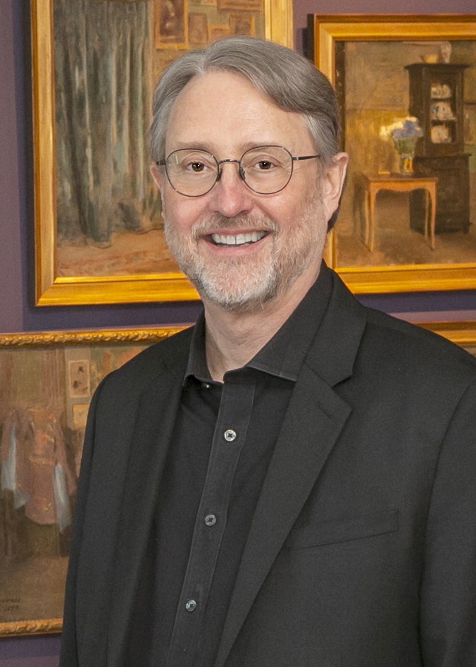 Man in black suit and shirt smiles in front of a painting