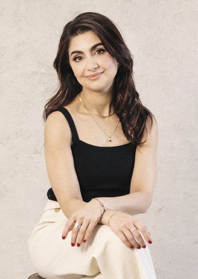 A woman with long brown hair wears a black top and white pants.