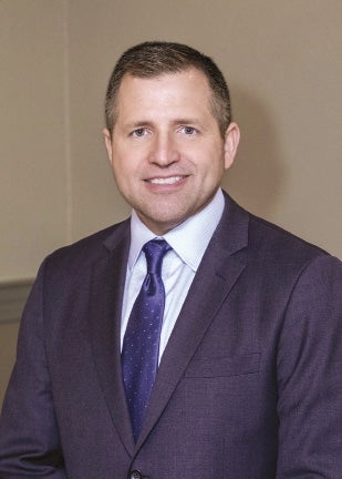 A man with short hair wears a dark blue suit jacket, white button down, and blue tie.