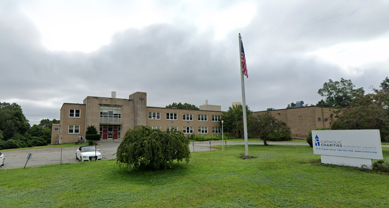 Three-story building sites behind small parking lot and a lawn with a flag and sign