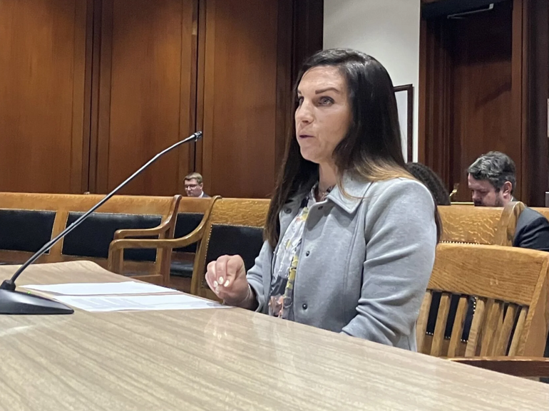Woman sits at table with microphone testifying at a hearing