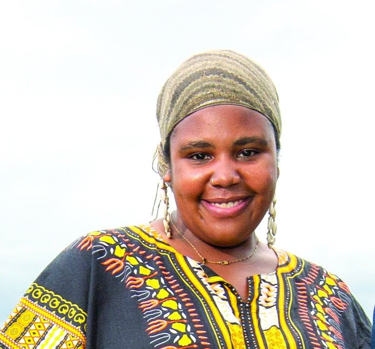 A woman wears a green patterned headwrap and a black, yellow, orange, and white patterned top.
