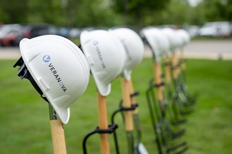 A line of eight shovels planted in the ground with white construction helmets labeled with Veranova's logo sitting on top of the shovels. 