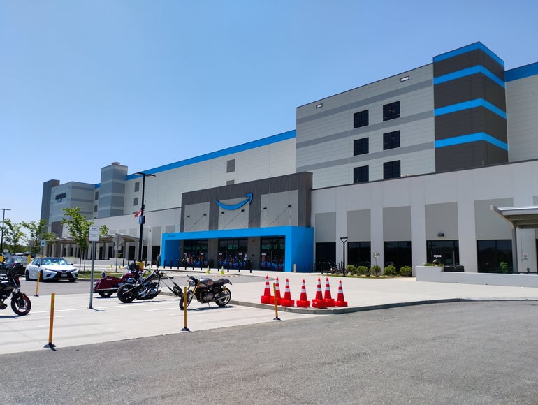 A large, five-story warehouse building sits behind a parking lot with cones and motorcyles in front of it. 