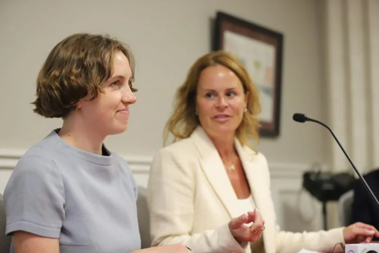 Two women sit at a table with a microphone in front of them. 