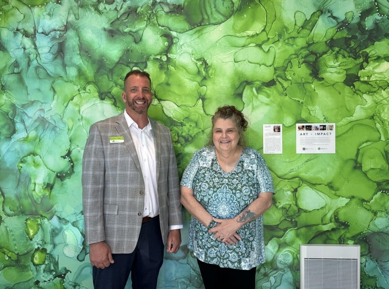 A man and a woman stand in front of a green wallcovering. 