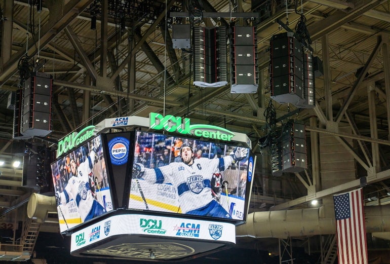 A stadium jumbotron with speakers next to it, all suspended from an arena ceiling