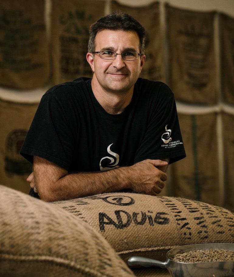 Man in black t-shirt leans on large bag of coffee beans