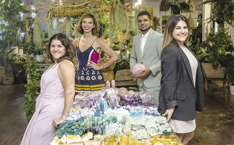 Four people stand inside a table full of crystals and gemstones inside a store. 